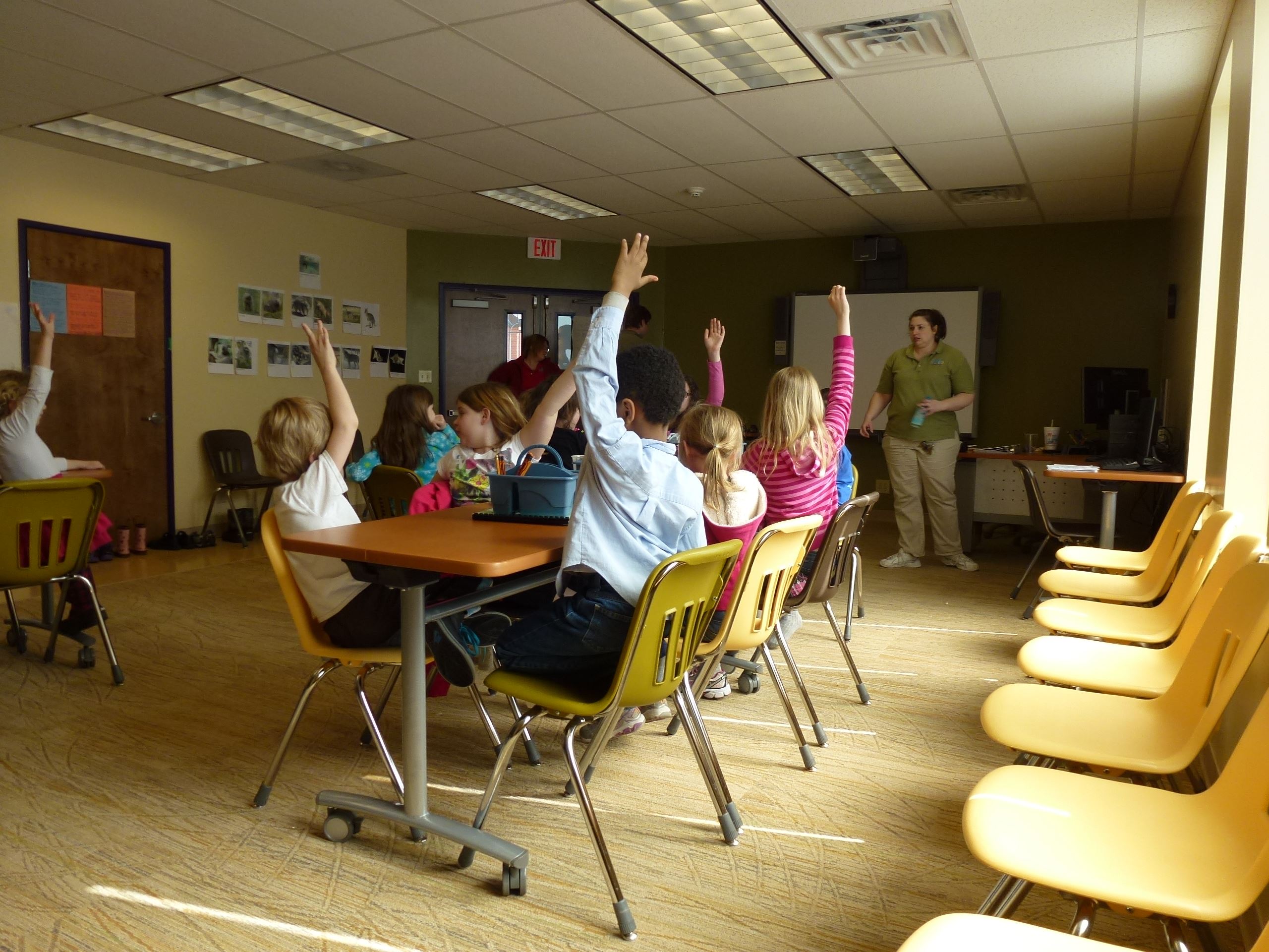 Kids seated at classroom tables raising hands to interact with the camp teacher