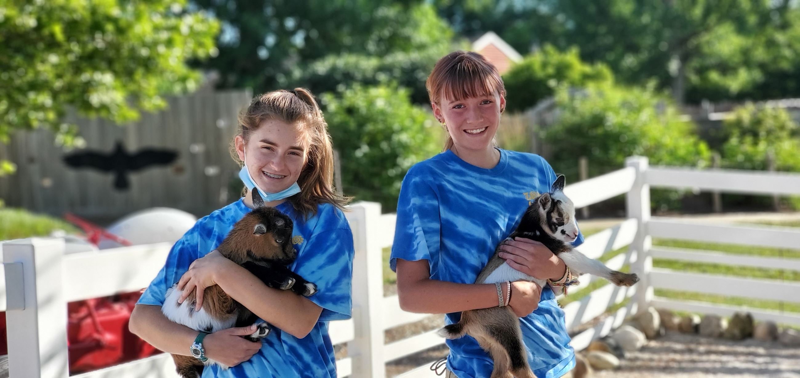 Two teenage girls wearing Zoo Teen uniform shirts pose holding baby goats